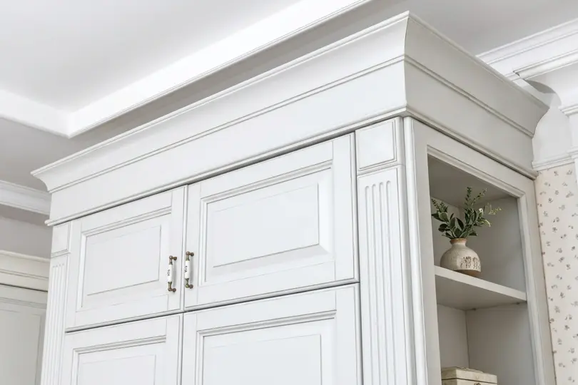 Detail of white cabinetry finished with layered crown molding, next to an arched glass-front display cabinet