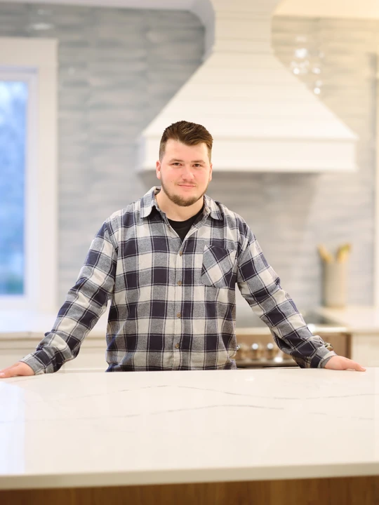 Blake Amarandos in a navy and grey flannel shirt standing behind a marble kitchen island with a white custom range hood behind him